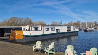 Houseboat De Zwerver attraccata a un molo di legno con sedie verdi sotto un cielo limpido.