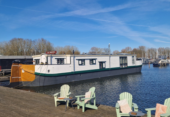 Casa flotante De Zwerver atracada en un muelle de madera con sillas verdes bajo cielo azul.