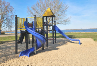 Spielplatz mit blauen Rutschen und gelbem Dach am Wasser, aufgenommen bei einer Glamping-Unterkunft.