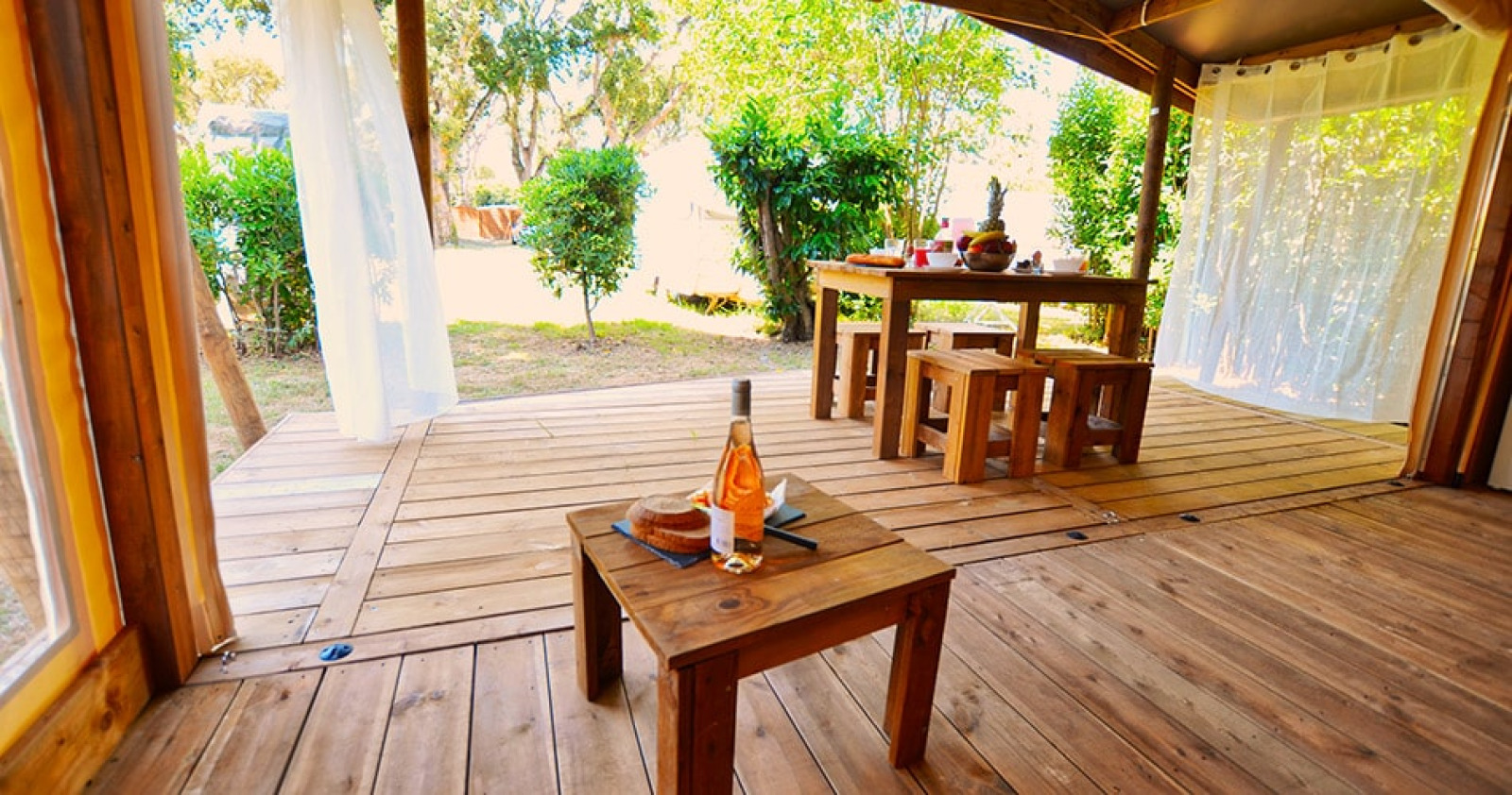 Houten terras met tafels en banken onder tentzeil bij Tent Lodge Riada, Camping Merendella in Frankrijk.