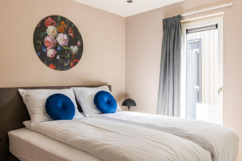 Bright bedroom with double bed, blue pillows, floral art, and window at Ecovilla Boszicht Comfort, Netherlands.