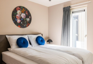 Dormitorio luminoso con cama doble, cojines azules, arte floral y ventana en Ecovilla Boszicht Comfort, Países Bajos.