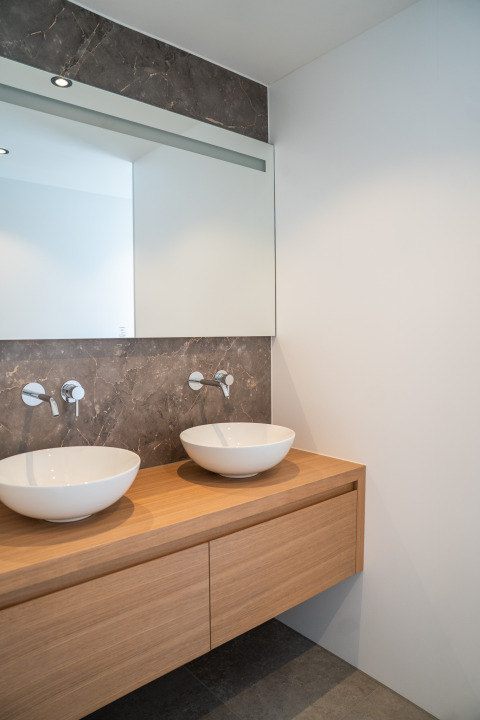 Moderno lavabo de baño con dos lavabos redondos sobre encimera de madera, espejo grande y pared de mármol.