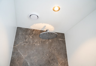 Modern showerhead in a lodge bathroom, featuring a marble wall, ceiling light, and ventilation fan.