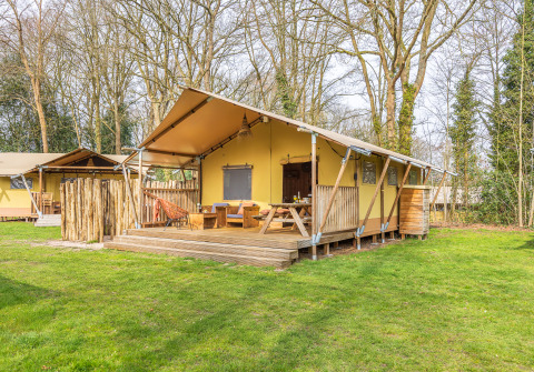Foto di una tenda safari con veranda in legno, sedute esterne e prato verde in un ambiente boschivo.