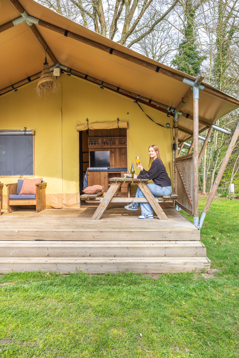 Kvinde nyder en drink ved et picnicbord foran safari-teltet 'De Bos Tent' på Gooise Heide, Holland.