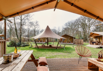 Udsigt fra en safaritelts terrasse med træmøbler, stole og store telte i en grøn, skovklædt campingplads.