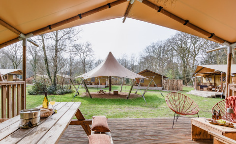 Vue d'une terrasse de tente safari avec mobilier en bois et grandes tentes sur un camping vert boisé.