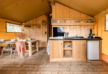 Interiør af et safaritelt med et træspisebord, køkkenområde, køleskab og åbne hylder med service.