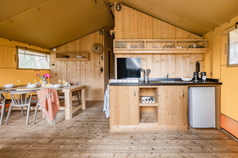 Interno di una tenda safari con tavolo da pranzo in legno, zona cucina, frigo e scaffali aperti.