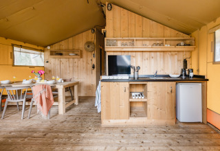Intérieur d'une tente safari avec une table en bois, une kitchenette, un petit frigo et des étagères.