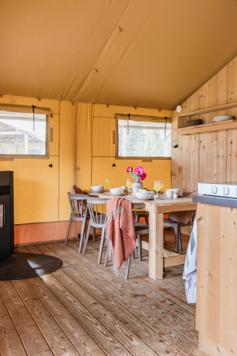 Interno di una tenda safari con tavolo in legno, sedie, stoviglie e fiori in un ambiente accogliente.