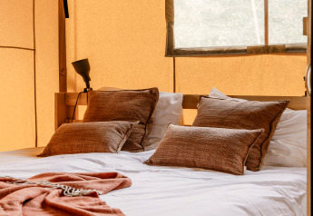 Komfortabel seng med brune puder og tæppe i en safariteltsuite med bløde beige vægge og naturligt lys.
