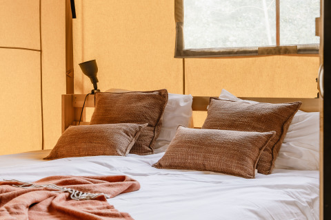 Lit douillet avec coussins bruns et couverture dans une tente safari chaleureusement éclairée par la lumière.