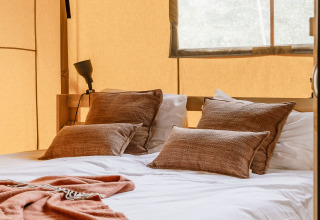 Lit douillet avec coussins bruns et couverture dans une tente safari chaleureusement éclairée par la lumière.
