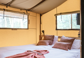 Intérieur d'une tente safari chic avec un grand lit, un plancher bois et de grandes fenêtres sur la nature.