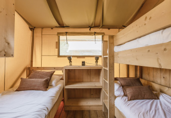 Intérieur d'une tente safari avec trois lits simples, une étagère en bois et une fenêtre éclairée.