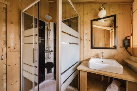 Salle de bain dans une tente safari avec cabine de douche, lavabo, miroir et murs en bois naturel.