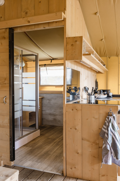 Interno di una tenda safari con pannelli in legno, angolo cottura e vista su doccia e zona pranzo.