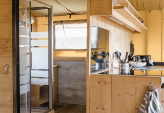 Intérieur d'une tente safari avec panneaux en bois, coin cuisine, et vue sur la douche et coin repas.