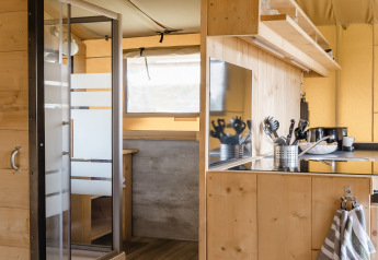 Intérieur d'une tente safari avec panneaux en bois, coin cuisine, et vue sur la douche et coin repas.