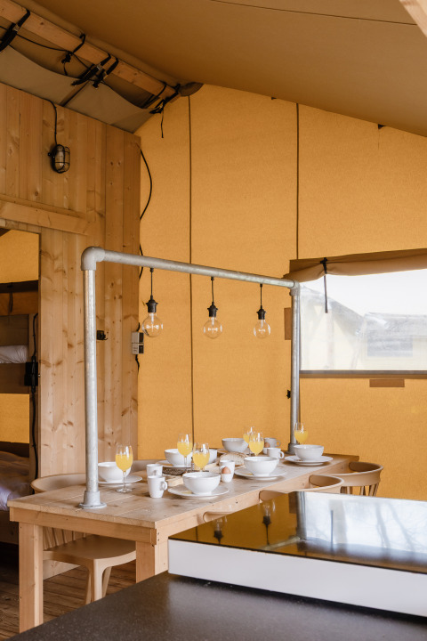 Spiseområde i et safaritelts interiør med træmøbler og dækket bord med morgenmad og juiceglas.