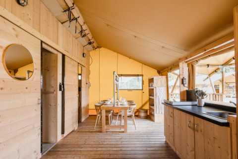 Vista interna di una tenda safari di lusso con arredi in legno, tavolo da pranzo e cucina aperta.