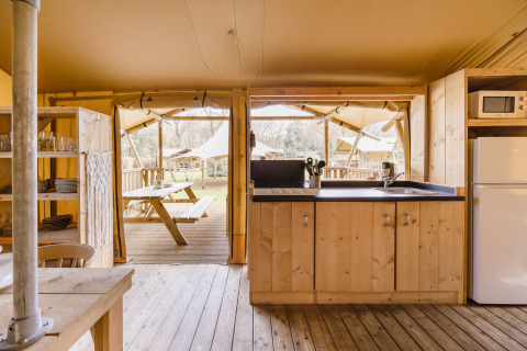Area cucina in una tenda safari con arredi in legno, vista su tavolo da picnic e altre tende all’esterno.