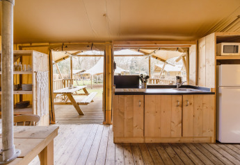 Keuken in een safaritent met houten inrichting, zicht op picknicktafel en andere tenten buiten.