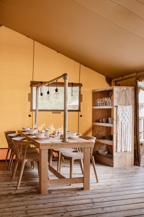 Zona pranzo in tenda safari con tavolo in legno, sedie, scaffale con stoviglie e illuminazione accogliente.