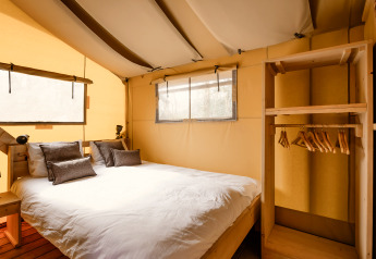 Innenraum eines Safarizeltes mit Doppelbett, Kissen, Holzregal und einer Stange mit Kleiderbügeln.