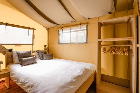 Interno di tenda safari con letto matrimoniale, cuscini, scaffale in legno, appendiabiti e luce naturale.