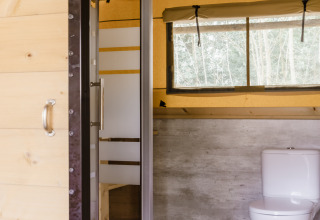 Bagno in tenda safari con doccia moderna, wc, pavimento in legno e ampia finestra sulla natura.