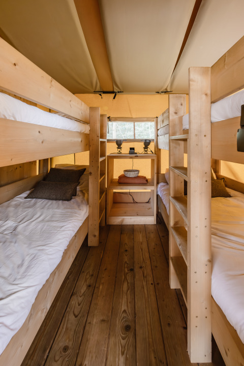 Vista interna di una tenda safari con pavimento in legno, letti a castello e una piccola mensola in fondo.