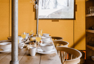 Tavolo da colazione con succo, ciotole e tazze in una tenda safari con sedie di legno e finestra.