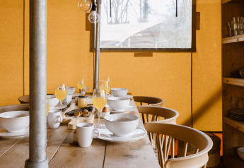 Ontbijttafel met fruitsap, kommen en kopjes in een safaritent met houten stoelen en raam.