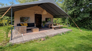 Tenda safari con veranda in legno coperta, panchine rustiche e ampio prato verde tra gli alberi.