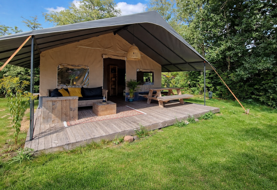Safari tent med stor overdækket terrasse, træmøbler og grønt græs, omgivet af frodige træer.