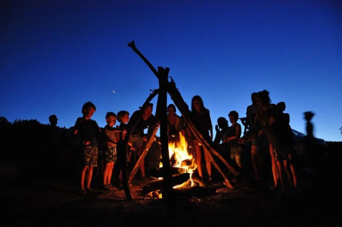 Kinder sitzen um ein Lagerfeuer bei Sonnenuntergang auf Agricampeggio Tasteria, einem Ferienpark in Marche, Italien.