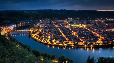 Luftfoto af en smuk, oplyst by ved en flod omkring Montcléra, Occitanie, Frankrig, taget ved skumring.