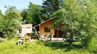Safari-Zelt inmitten von grünen Bäumen und hohem Gras an einem sonnigen Tag mit Holzmöbeln davor.