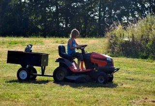 Barn kører på en plænetraktor med en trailer i en solrig park ved Les Savonniers i Occitanie, Frankrig.
