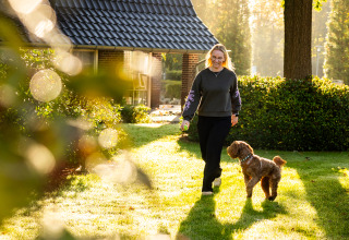 Vrouw wandelt met hond bij Wellness Lodge XXL op Hof van Salland in Nederland, omringd door zonlicht.