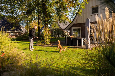 Hombre juega con perro en el jardín soleado de Wellness Lodge XL en Hof van Salland, Países Bajos, con casa al fondo.