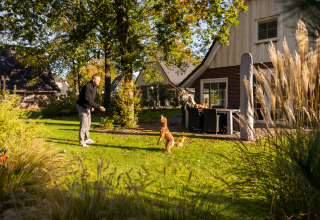 Hombre juega con perro en el jardín soleado de Wellness Lodge XL en Hof van Salland, Países Bajos, con casa al fondo.