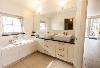 Baño luminoso con dos lavabos, bañera con espuma y gran espejo en Wellness Lodge XL, Hof van Salland, Países Bajos.