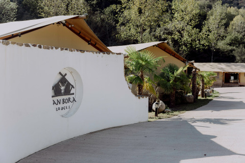 Ingang van Can Bora Lodges vakantiepark in Catalonië, Spanje, met tenten, palmbomen en witte muur.