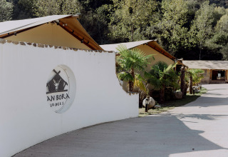 Ingang van Can Bora Lodges vakantiepark in Catalonië, Spanje, met tenten, palmbomen en witte muur.