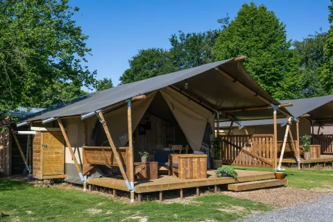 Vista esterna di una tenda safari Glampingtent 4p con veranda in legno e sedie, circondata da alberi.