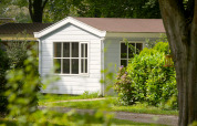 Wit chalet op Camping Mareveld in Nederland, omgeven door groene struiken en bomen in de zon.